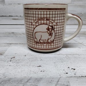 Puede incluir: Taza de cerámica blanca con un patrón de cuadrícula marrón y una ilustración de vaca marrón. La taza tiene las palabras "Country Best" impresas.