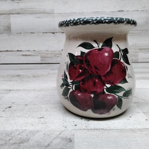 May include: A cream-colored ceramic jar with a decorative apple design. The jar features a cluster of red apples with green leaves. The rim is decorated with a green and white pattern. This kitchenware item is suitable for storing utensils or as a decorative piece.