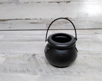 Op de afbeelding: Een zwarte ketel met een gebogen handvat, ideaal voor Halloween of thema-decoratie. De ketel heeft een ronde body met decoratieve bladerpatronen en een brede opening. Het handvat is van zwart metaal.