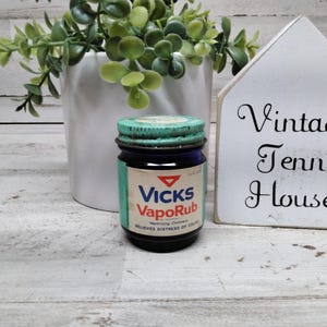Könnte beinhalten: Ein blaues Vintage-Glasgefäß von Vicks VapoRub mit einem verwitterten, türkisfarbenen Metallschraubdeckel und einem weißen Papieretikett, das das rote Dreieckslogo, den Text Vicks VapoRub und den Schriftzug Relieves Distress of Colds zeigt.
