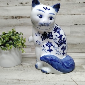 Op de afbeelding: Een witte keramische kattenfiguur met blauwe bloempatronen en accenten. De kat heeft blauwe ogen, oren en een staart. Links van de kat staat een kleine potplant en rechts een witte keramische haan.