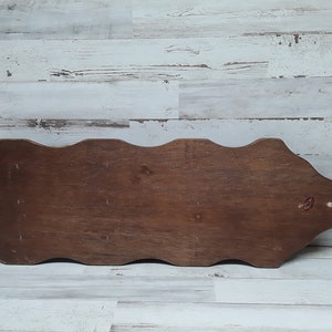 May include: A wooden decorative wall shelf with a wavy top edge and a dark brown finish. The shelf has a rectangular shape with a rounded end and a small red sticker. The background is a white and gray wooden wall.