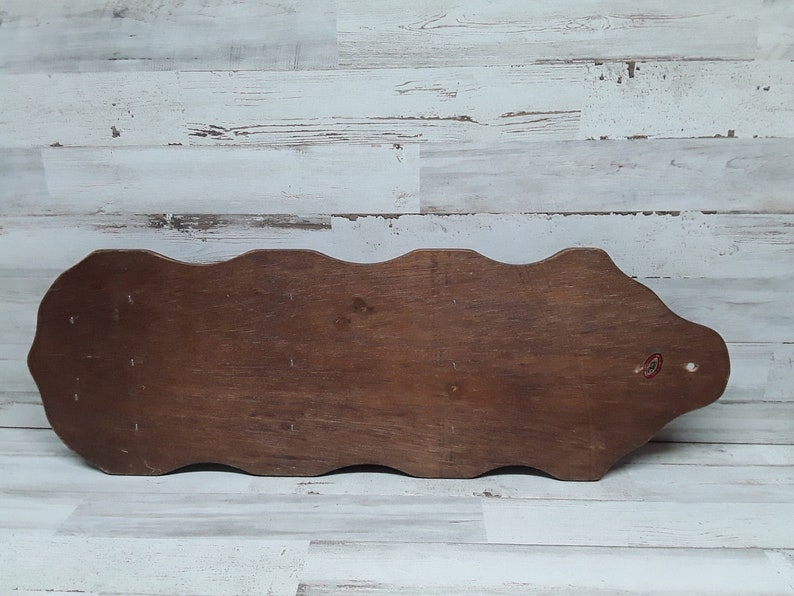 May include: A wooden decorative wall shelf with a wavy top edge and a dark brown finish. The shelf has a rectangular shape with a rounded end and a small red sticker. The background is a white and gray wooden wall.
