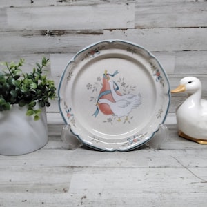 Puede incluir: Un plato decorativo con borde festoneado y ribete azul. El plato presenta un diseño de dos gansos blancos con una cinta roja y azul. Un pato de cerámica blanco y una pequeña planta en maceta también están en la imagen.