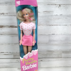 May include: A vintage Barbie doll in its original packaging. The doll has blonde hair and wears a pink skirt and a white top with pink hearts. The box is pink and features the text "Pretty Hearts Barbie".
