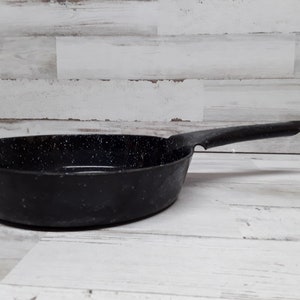 Puede incluir: Una sartén de hierro fundido negro moteado con un mango largo. La sartén es redonda con un borde ligeramente ensanchado. El interior tiene un patrón moteado. El mango está unido al lateral de la sartén.