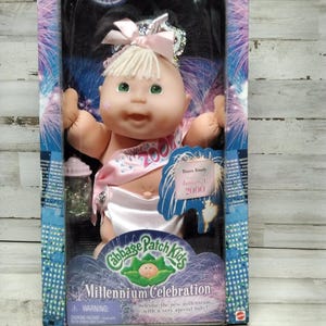 Puede incluir: Muñeca Cabbage Patch Kids Millennium Celebration en su embalaje original. La muñeca tiene cabello rubio, ojos verdes y una banda rosa. La caja presenta un diseño azul y rosa con el texto "Millennium Celebration".