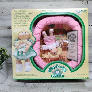 Puede incluir: Un juego de muñecas Cabbage Patch Kids Pin-Ups vintage en su caja original. La caja presenta una muñeca que sostiene una muñeca más pequeña y otra muñeca dentro de un marco rosa. La caja tiene el texto "Cabbage Patch Kids" y "Pin-Ups".