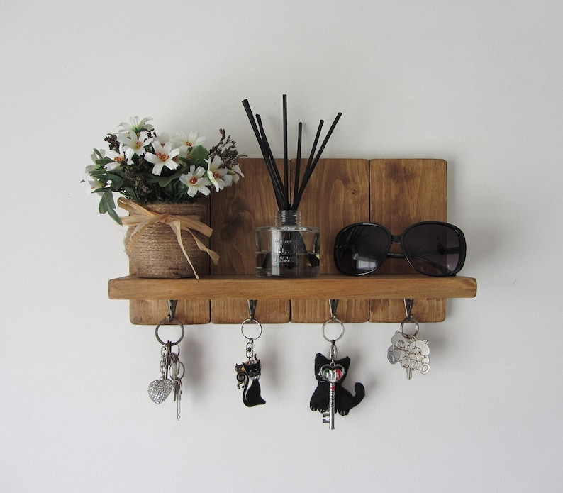 May include: Wooden key holder with a shelf, five hooks, and a small potted plant. There are sunglasses, a diffuser, and keys hanging on the hooks.