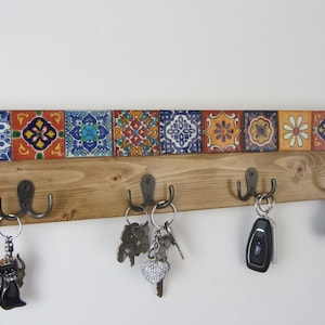 Puede incluir: Un perchero de llaves de madera con cinco ganchos y un borde de azulejos decorativos. Los azulejos son coloridos y presentan patrones geométricos en tonos de azul, naranja, amarillo y verde. El perchero está montado en una pared blanca y tiene llaves colgando de los ganchos.
