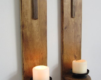 Könnte beinhalten: Zwei Wandleuchter aus Holz mit Kreuzdesign und Kerzenhaltern. Jeder Leuchter hat ein braunes Holzkreuz und einen schwarzen Kerzenhalter aus Metall. Die Leuchter sind aus Naturholz gefertigt und haben einen rustikalen Look.