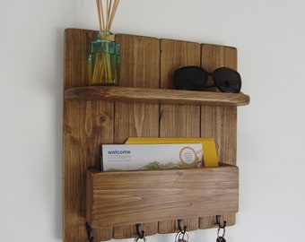 Peut inclure: Porte-clés mural en bois rustique avec une étagère, une fente à courrier et cinq crochets. Le porte-clés est teinté d'une couleur brun foncé. Trois porte-clés sont accrochés aux crochets et une paire de lunettes de soleil est sur l'étagère. Un petit flacon de diffuseur de roseaux se trouve sur l'étagère.