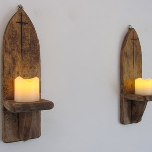 Könnte beinhalten: Zwei Wandleuchter aus Holz mit einem Kreuzdesign und einem Regal für eine Kerze. Jeder Leuchter hat eine brennende Kerze auf dem Regal.