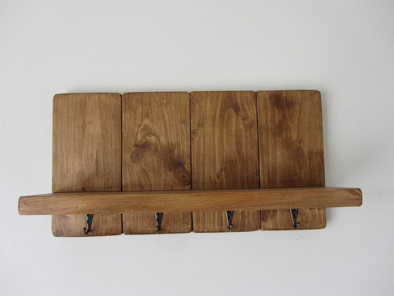 May include: A rustic wooden shelf with four hooks, stained in a dark brown color. The shelf is made of reclaimed wood and has a natural, distressed look.