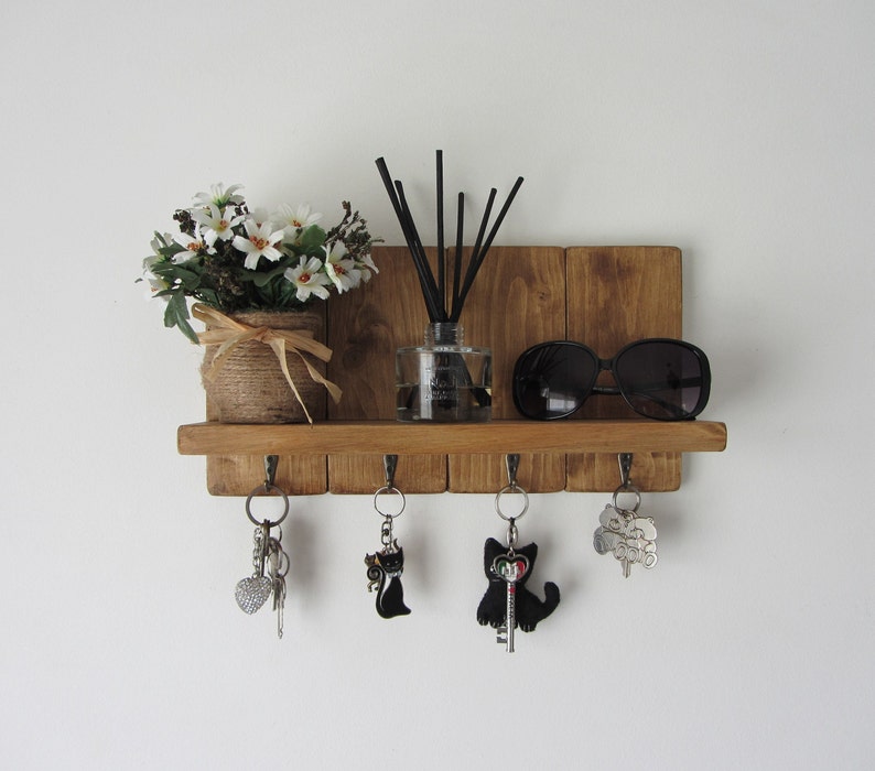 May include: Wooden key holder with five hooks, a shelf, and a small potted plant. There are also sunglasses and a diffuser with reeds on the shelf.