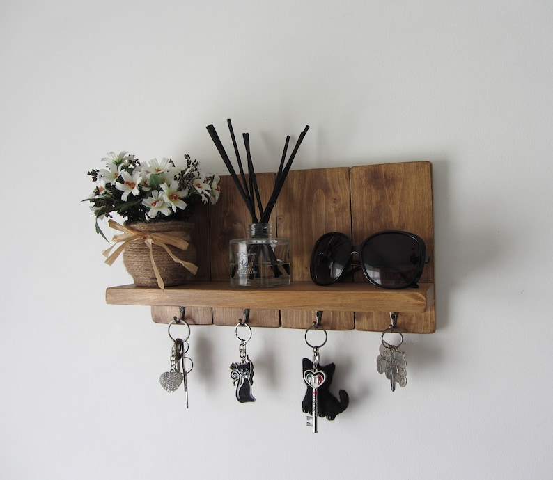 May include: A wooden key holder with a shelf and five hooks. The shelf has a small potted plant, a bottle of reed diffuser, and a pair of sunglasses. The hooks have keys with cat-shaped charms.