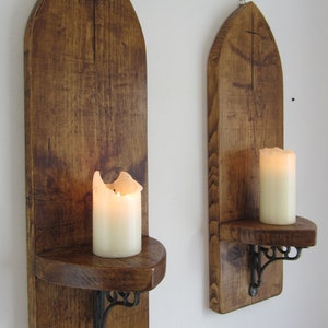 Puede incluir: Dos apliques de pared de madera con un diseño de cruz y un estante para velas. Cada aplique tiene un soporte de metal negro y un estante para una vela. Los apliques están hechos de madera marrón oscura y tienen un aspecto rústico.