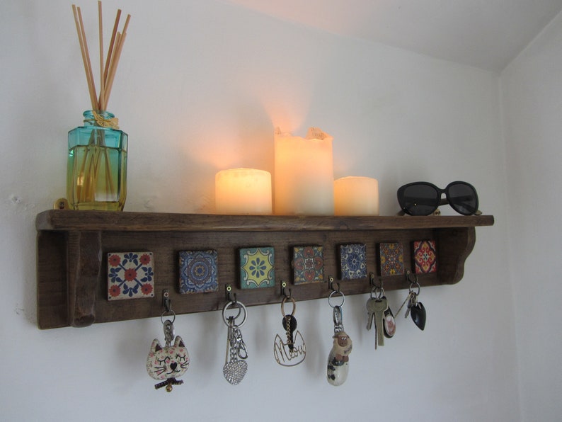 May include: A wooden key rack with six hooks and colorful tile accents. The rack is mounted on a white wall and has candles and a diffuser on the shelf above.