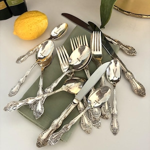 May include: An assortment of antique silverware, including spoons, forks, and knives, with ornate floral detailing. The silverware is arranged on a green napkin, with a lemon and a few bottles in the background.