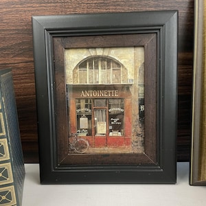 May include: A framed print featuring a Parisian restaurant scene. The print, set in a black frame, depicts a red-fronted building with the name "ANTOINETTE" above the entrance. A bicycle is parked outside.