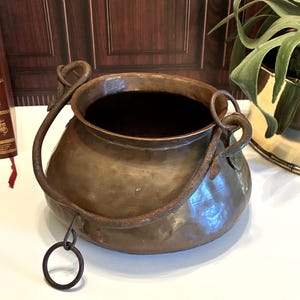 May include: A round, hammered copper pot with a wide opening and a dark, weathered iron swing handle. The handle features decorative, twisted metal loops at the attachment points and a small circular ring hanging from the bottom of the handle.