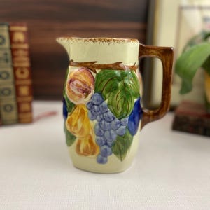 May include: Cream-colored ceramic pitcher with a brown handle and spout. The pitcher is decorated with colorful grapes, leaves, and fruit. The handle and rim have a brown trim. The pitcher is sitting on a white surface.