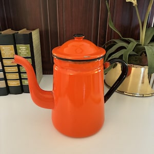 May include: A bright orange enamel coffee pot with a black handle, a matching lid with a black knob, and a long curved spout. The pot features a smooth, glossy finish with a black rim around the top edge and a small chip on the spout tip.