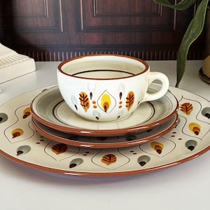 May include: A ceramic dinnerware set consisting of a cup, a saucer, and a plate, all featuring a cream-colored base with a repeating pattern of stylized orange, yellow, and grey leaf-like motifs and a solid brown rim on each piece.
