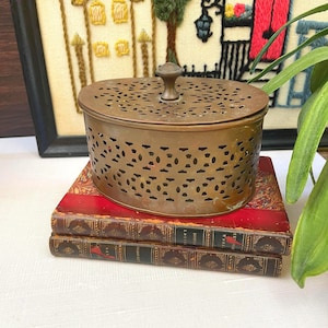 May include: A vintage, round, bronze-colored metal container with a perforated design and a small knob on top. The container sits atop two antique books with red and gold covers, against a backdrop of framed embroidery.