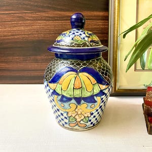 May include: A ceramic jar with a lid, featuring a vibrant hand-painted design. The jar showcases a blue, yellow, and green floral pattern on a white checkered background. The lid and finial are a deep blue, adding to the decorative appeal.