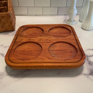 May include: A square wooden serving tray with four circular indentations. The tray is made of reddish-brown wood and has a raised edge. The tray is designed to hold four glasses or mugs.