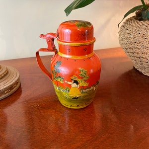 May include: A vintage, orange-red metal pitcher with a hinged lid and handle. The pitcher is decorated with a painted scene of a girl in a yellow dress and hat in a field. The pitcher has yellow and green accents.