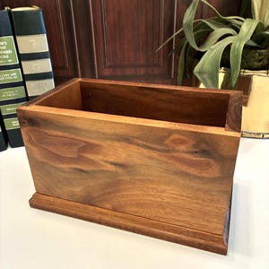 May include: A rectangular wooden box with a natural brown wood grain finish. The container features a flat base and four vertical sides forming an open top. The wood displays varied light and dark brown tones throughout the surface of the structure.