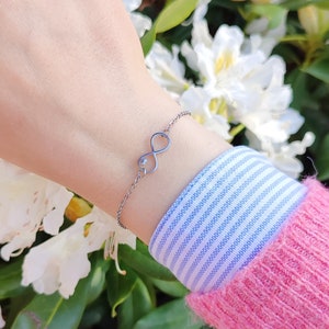 Könnte beinhalten: Ein silberfarbenes Metallkettenarmband mit einem zentralen Unendlichkeitssymbol als Anhänger. Das Unendlichkeitssymbol ist offen gestaltet und weist auf einer Seite der Schlaufe ein kleines, filigranes, herzförmiges Ausschnittdetail auf.
