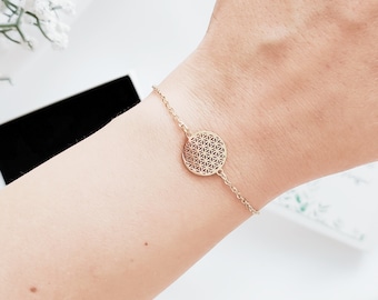 May include: A gold bracelet with a fine chain and a circular pendant. The pendant has a geometric pattern. The bracelet is shown on a wrist, emphasizing its elegant and minimalist design. The bracelet is a stylish piece of jewellery.