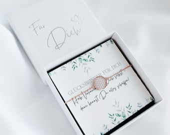 May include: A rose gold bracelet with a geometric pattern, displayed in an open white gift box. The box contains a card with text and botanical illustrations. The box lid is inscribed with the words "Für Dich".