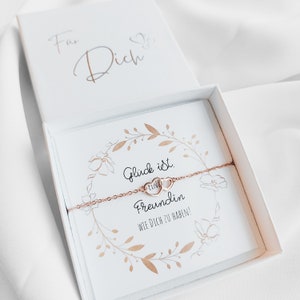 Könnte beinhalten: Ein roségoldenes Armband mit zwei herzförmigen Anhängern, präsentiert in einer offenen weißen Geschenkbox. Die Box enthält eine Karte mit dem Schriftzug "Glück ist Freundin" und einem floralen Design. Die Box liegt auf weißem Stoff.