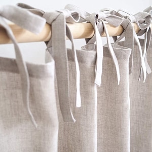 Puede incluir: Conjunto de seis cortinas de lino beige con lazos blancos, colgadas de una barra de madera.