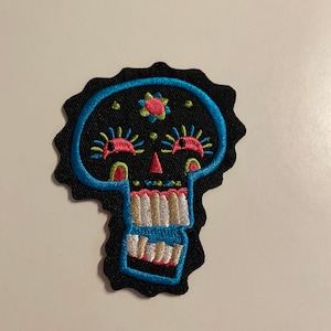 Puede incluir: Parche bordado de calavera con fondo negro, contorneado en azul. La calavera presenta detalles en rosa, amarillo y blanco, incluyendo una flor en la frente y detalles decorativos de ojos y dientes. El parche tiene un borde festoneado.