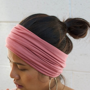 May include: A dusty rose-colored wide headband is wrapped around the head. The headband is made of a soft, stretchy material. The person is wearing gold hoop earrings. The hair is pulled back in a bun.