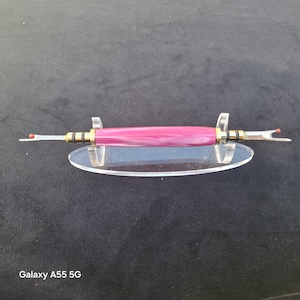 May include: A double-ended seam ripper featuring a polished magenta and white marbled barrel with gold-tone metal accents. The tool has two silver-coloured metal blades with small red safety balls at the tips, resting on a clear acrylic display stand.