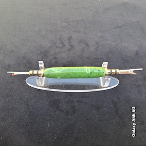 May include: A seam ripper featuring a green, speckled, cylindrical handle with light green swirl patterns. The tool has gold-toned metal accents and two silver-coloured metal prongs with small red safety balls at the tips, resting on a clear stand.