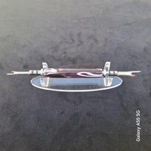 May include: A double-ended seam ripper featuring a central barrel with a dark purple and white marbled resin pattern. The tool has metallic silver-toned hardware and two sharp metal blades with small red safety balls at the tips, resting on a clear stand.