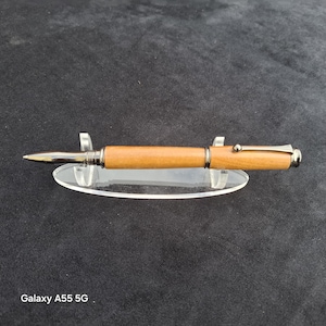 May include: A ballpoint pen with a light brown wooden barrel and polished gunmetal hardware, including a pointed tip, a centre band, and a pocket clip, resting horizontally on a clear, oval-shaped acrylic display stand.