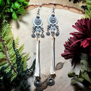 May include: A pair of long dangle earrings featuring silver-toned metal components with circular, textured patterns at the top. Each earring includes a long, slender, off-white bone segment suspended vertically, ending in a small, circular metal charm.