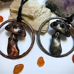 Puede incluir: Dos collares con colgantes de setas. Un colgante es de color cobre, el otro es plateado. Cada seta está dentro de un marco circular y cuelga de un cordón negro. Hay piedras naranjas dispersas alrededor de los colgantes. El fondo incluye piedras blancas y grises.