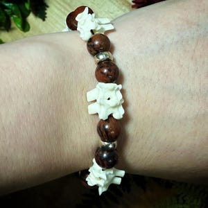May include: A stretch bracelet featuring alternating white bone-like vertebrae segments and polished dark brown spherical beads, accented with small metallic silver-toned spacer beads arranged in a repeating pattern around the circular band.