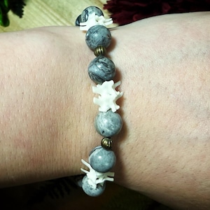 May include: A stretch bracelet featuring alternating round grey marbled stone beads and white bone-shaped spacers, accented with small metallic bronze-toned ribbed beads. The beads are arranged in a repeating pattern along the elastic cord.