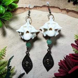 May include: A pair of dangle earrings featuring white animal vertebrae bones suspended from metal hooks. Each earring includes a small, round, light green bead and a dark bronze-toned metal pendant shaped like a pointed leaf with etched details.