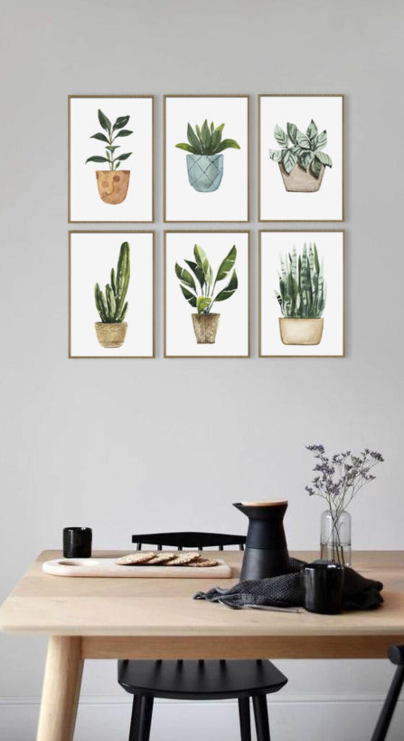 May include: A set of six watercolor prints featuring different potted plants. The prints are framed in a simple gold frame and are arranged in a grid pattern on a white wall.